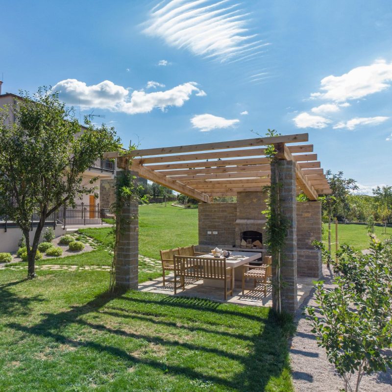 A wide shot of a wood and stone building next to a wooden pergola in a garden.