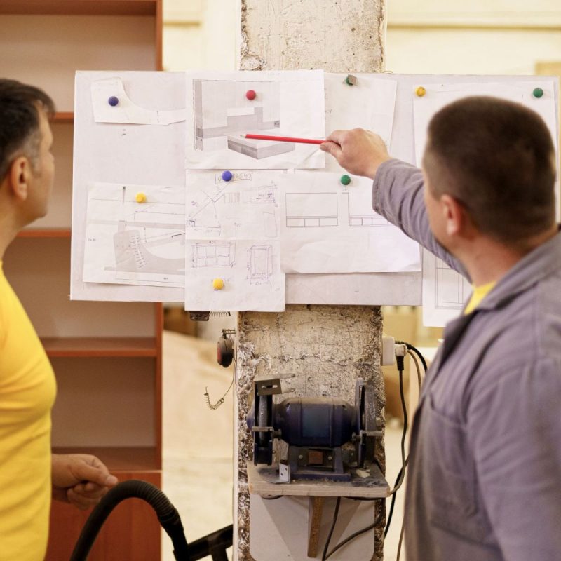 Carpenters working on furniture projects Two colleagues carpenters discussing and working with a technical blueprint drawing construction paper in their workshop illustrating models on a board