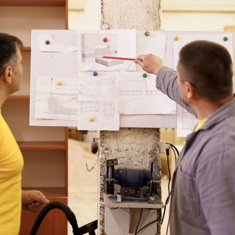 Two colleagues carpenters discussing and working with a technical blueprint drawing construction paper in their workshop illustrating models on a board