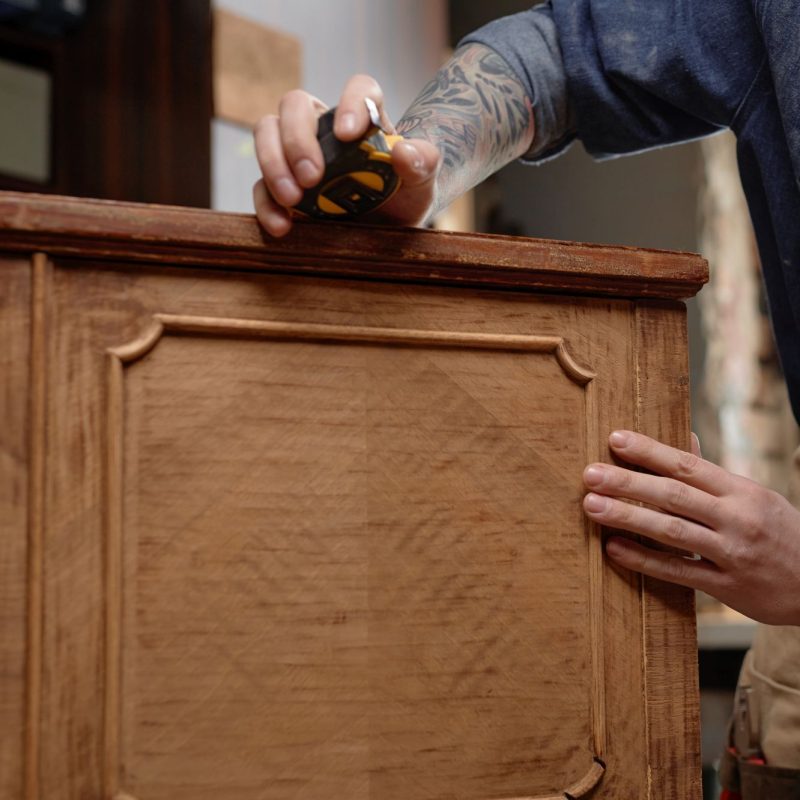 Carpenter with tattooed arms working on woodworking project, using tape measure on wooden surface in workshop environment with detailed focus on hands while measuring